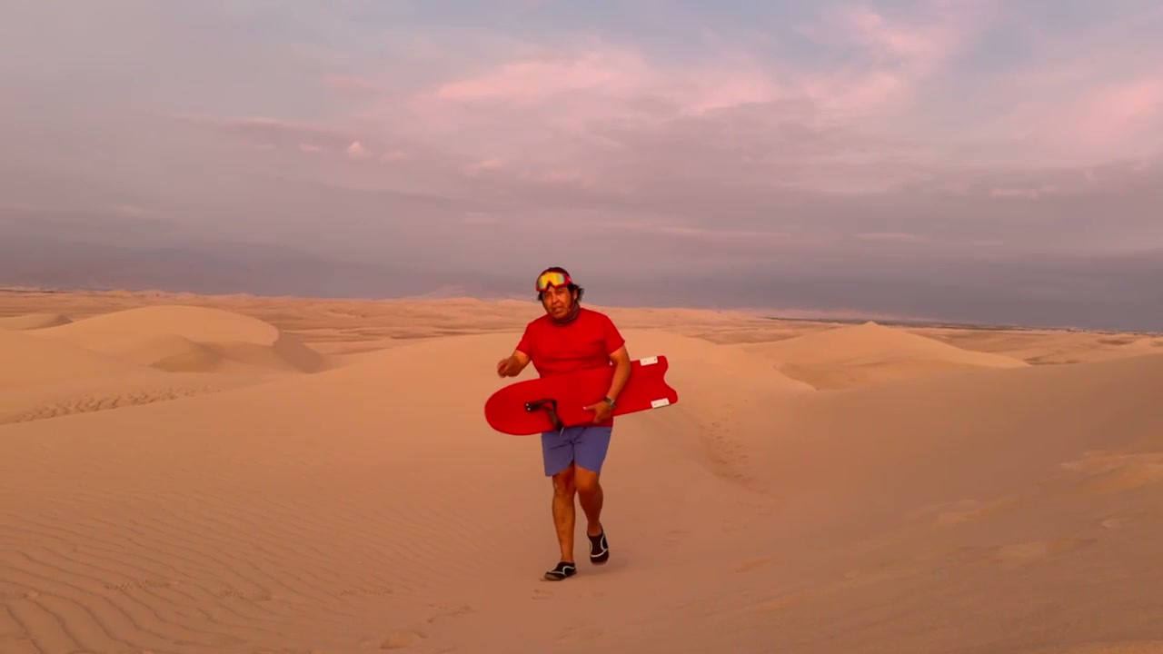 Hombre camina por la cresta de una duna al atardecer con una tabla de sandboarding roja